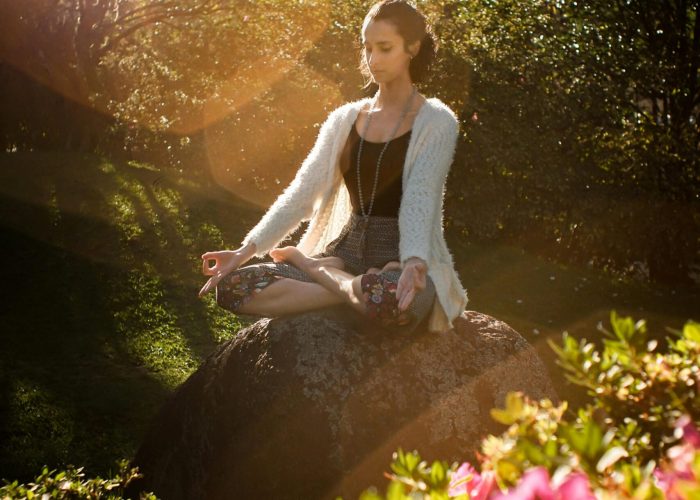 Woman in tranquil meditation pose outdoors, bathed in sunlight, surrounded by lush greenery.